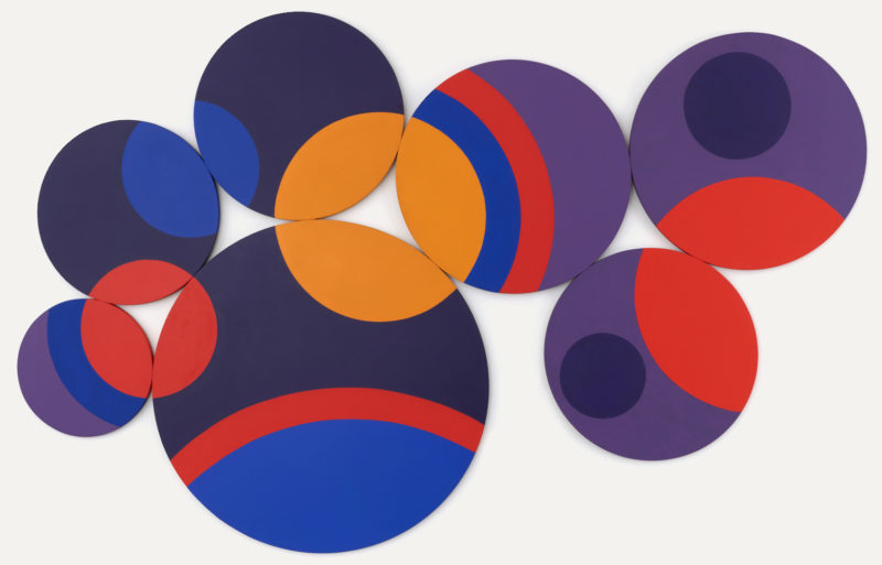 A geometric composition comprised of seven circular canvases of various sizes. They seem attracted touching as if bound together to form a single 'constellation'. Within the canvases are more circles, some are large spanning multiple canvases. Bright colors bring this work to life, the color scheme can be described as energetic with primary blue and red alongside a few shades of purple. Accenting the center of the work is an orangeish yellow circle.